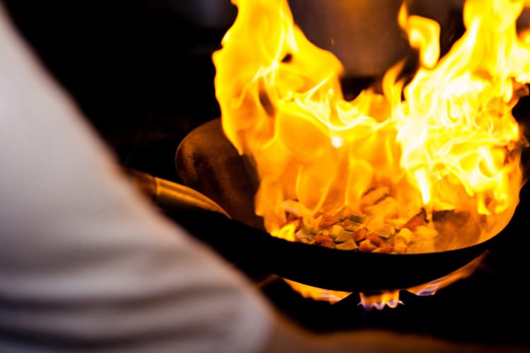 Llama de fuego flambeando y salteando verduras en sartén sobre fogón de gas.