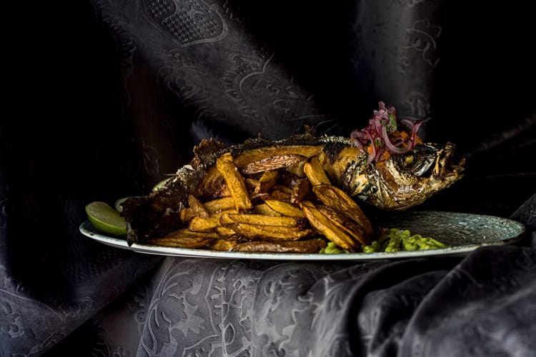 Plato de lubina frita sobre mousse de aguacate y acompañada de patatas fritas y limón.