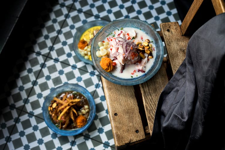 Chef con cuchara emplatando dos platos de causa peruana sobre mesa de madera oscura.