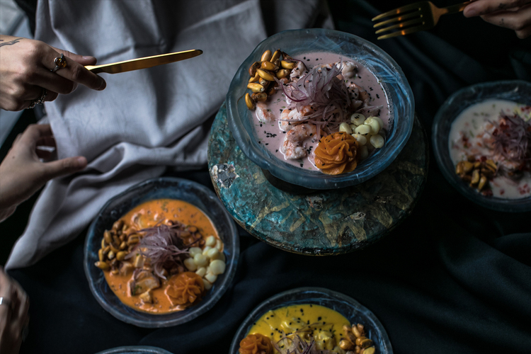 Mano con cuchara emplatando dos platos de causa peruana sobre mesa de madera.