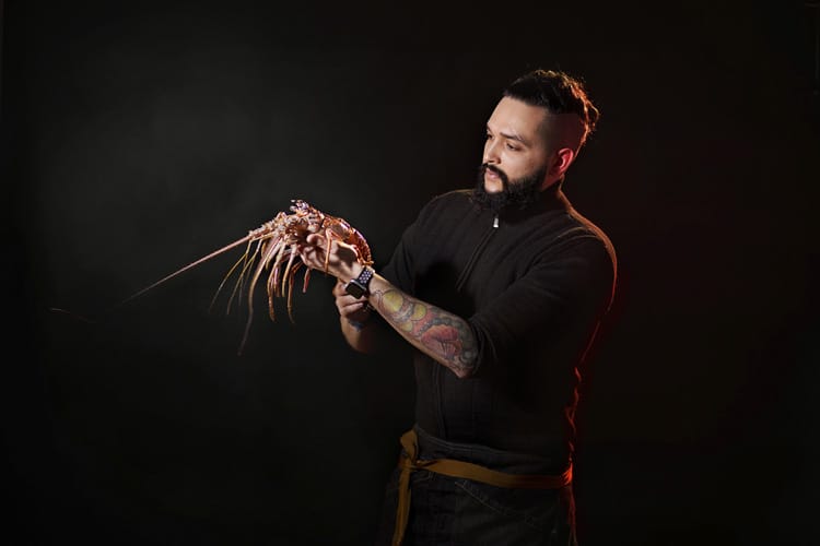 Chef hombre con barba tatuado sujetando una langosta cruda.