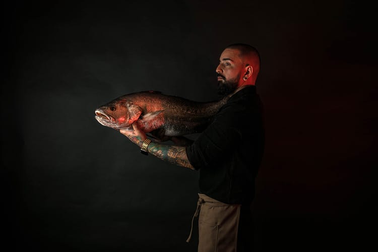 Chef hombre con barba tatuado sujetando pescado crudo.
